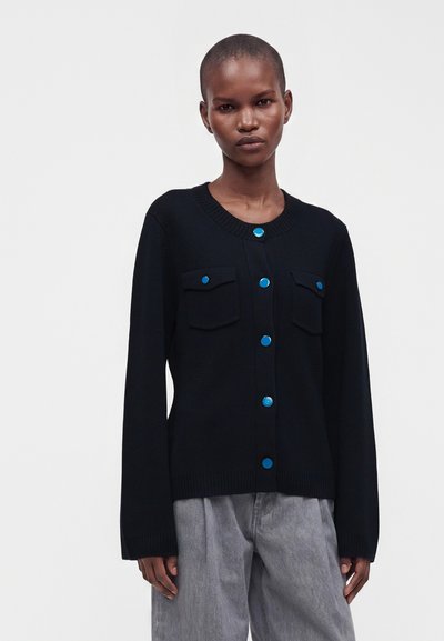 Cardigan noir boutonné avec des boutons bleus et des poches porté sur un pantalon gris par une personne aux cheveux courts sur un fond uni.