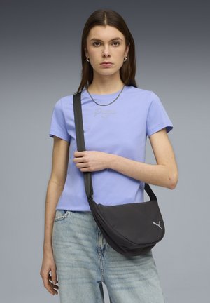 Zwart crossbodytasje met een gebogen vorm en een Puma-logo. Model draagt een lichtpaars T-shirt met ronde hals en casual blauwe spijkerbroek.