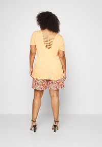 Žluté tričko s hlubokým výstřihem do V na zádech a proplétanými detaily, doplněné květovanými šortkami, které zdůrazňují živé barvy a vzory. Černé podpatky.