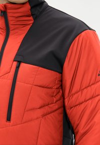 Veste rouge et noire avec un design matelassé, dotée d'un col haut, d'une poche poitrine zippée et d'une texture de tissu lisse.