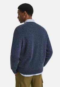Mann in einem blauen Strickpullover über einem blauen gestreiften Hemd, trägt olivgrüne Hosen und steht mit beiden Händen in den Taschen nach links gewandt.