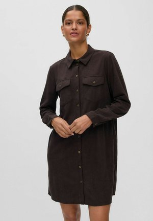 Femme portant une robe chemise marron foncé à boutons, manches longues et deux poches poitrine, debout devant un fond clair uni.