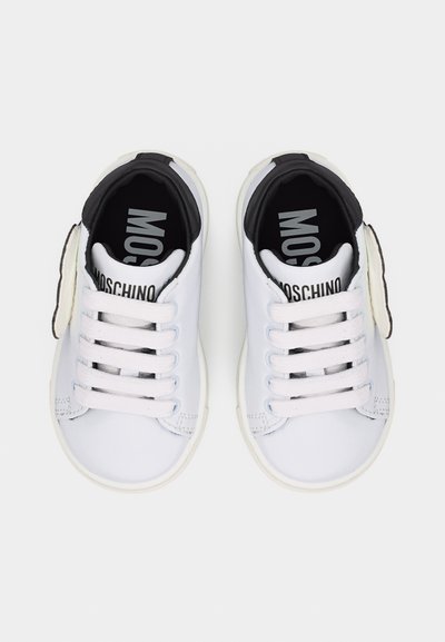 Λευκά παιδικά αθλητικά παπούτσια με μαύρη εσωτερική επένδυση, λευκά κορδόνια και το λογότυπο "MOSCHINO" στη γλώσσα και την πάτο, απεικονιζόμενα από πάνω.