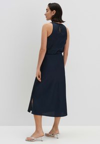Marineblaues ärmelloses Kleid mit tailliertem Schnitt, A-Linien-Silhouette, Schlüssellochausschnitt hinten und seitlichen Schlitzen, kombiniert mit silbernen Absatzsandalen.