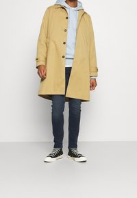 Beige sadetakki nappeilla ja hupulla, joka on puettu vaaleansinisen hupparin ja tummien farkkujen päälle. Mustavalkoiset tennarit täydentävät asun.