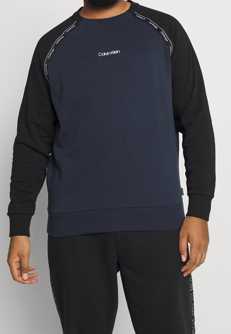 Sudadera azul marino oscura con mangas raglán negras, puños y dobladillo acanalados. Presenta un logo de Calvin Klein en el pecho y una marca sutil en las mangas.