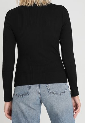 Person trägt ein eng anliegendes schwarzes Langarmoberteil und hellblaue Jeans, von hinten vor einem einfarbigen Hintergrund gezeigt.