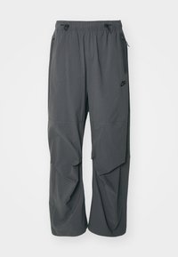 Pelēkie Nike bikses ar brīvu piegriezumu, kurām ir elastīga josta, aukla un kabatas ar rāvējslēdzējiem sānos. Izgatavotas no viegla auduma ar gludu tekstūru.