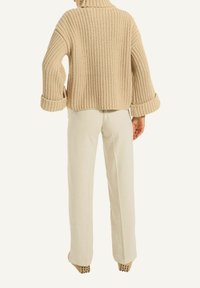 Beige Strickpullover mit hohem Kragen und lockeren Ärmeln, kombiniert mit cremefarbenen weit geschnittenen Hosen und tanfarbigen Absatzschuhen mit Nietenakzenten.