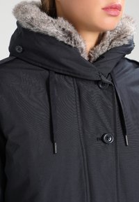 Czarna kurtka wierzchnia z kapturem podszytym sztucznym futrem, zapinana na guziki z ściągaczem. Gładka faktura materiału z subtelnymi detalami projektowymi.