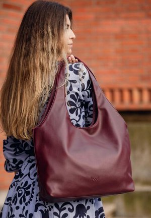 Kastanienbraune Leder-Hobo-Tasche mit glatter Textur, großem Öffnung und einfachem Schnitt, gehalten von einer Person, die ein Kleid mit Blumenmuster trägt.