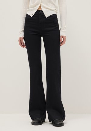 Persona que lleva pantalones negros acampanados de talle alto, zapatos negros y una camiseta blanca de manga larga con botones en la cintura.