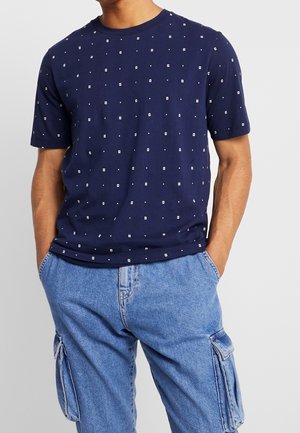 Navy-blaue Baumwoll-T-Shirt mit Rundhalsausschnitt, das ein geometrisches Muster aus weißen Quadraten und Punkten aufweist. Kurze Ärmel und lockere Passform.