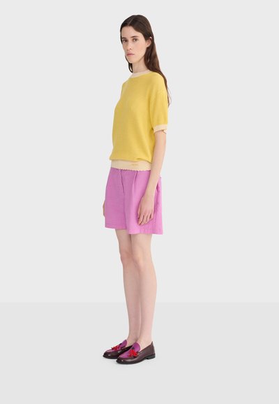 Femme debout portant un pull jaune à manches courtes, un short rose et des mocassins brillants violet foncé avec des glands rouges, sur un fond clair uni.