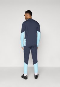 Άνδρας που φοράει ναυτικό και ανοιχτό μπλε αθλητικό κοστούμι, στέκεται μπροστά σε έναν απλό ανοιχτό γκρίζο τοίχο, με μαύρα παπούτσια και λευκές κάλτσες ορατές.