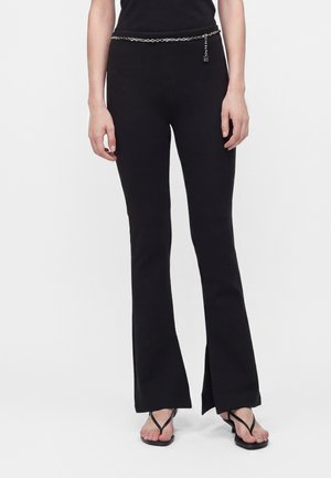 Personne portant un pantalon noir évasé taille haute avec des fentes latérales, une ceinture chaîne et des sandales noires, debout devant un fond uni.