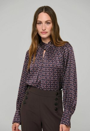 Donna con lunghi capelli castani che indossa una camicetta fantasia con collo annodato e pantaloni scuri a vita alta con bottoni ai lati.