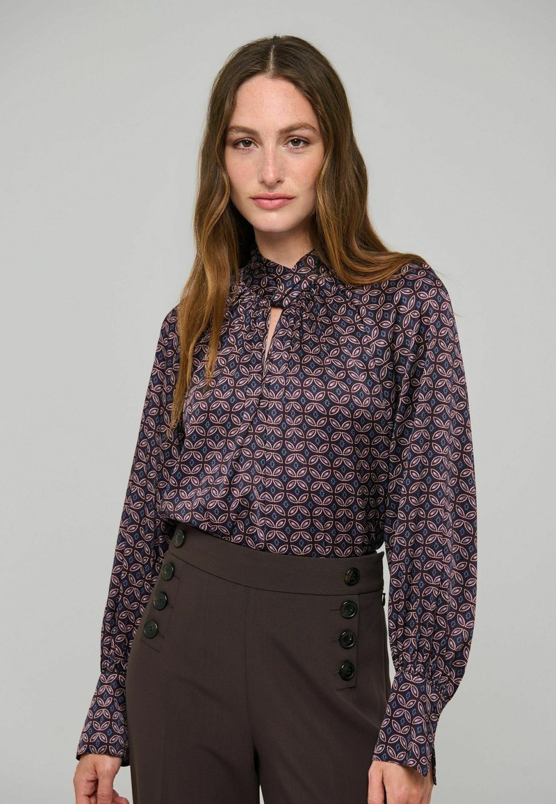 Femme aux longs cheveux bruns portant un chemisier à motifs avec un col noué et un pantalon taille haute foncé avec des boutons sur les côtés.