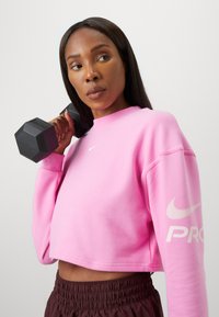 Różowy skrócony sweatshirt z białym logo Nike na piersi i rękawie, dopasowany do brązowych spodni na elastycznej talii, trzymający sześcienny czarny ciężarek.