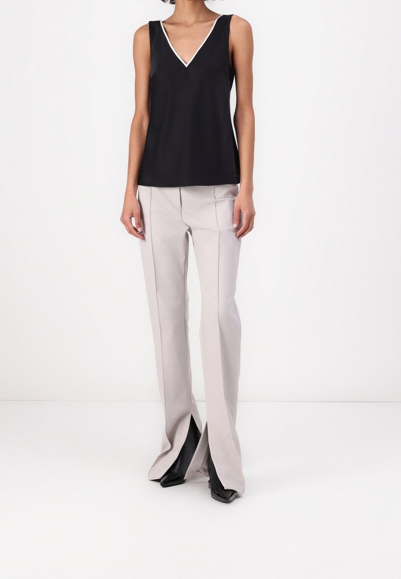 Femme portant un haut noir sans manches à col en V avec bordure blanche, un pantalon large gris clair avec des fentes à l'avant, et des chaussures noires à bout pointu.