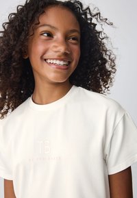 Tricou crem deschis cu mâneci scurte, cu un logo subtil brodat "TB" în roz și textul "by TED BAKER" dedesubt.