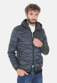 Uomo con barba che indossa una giacca imbottita nera con cappuccio sopra una camicia bianca e jeans blu, che tiene la cerniera della giacca vicino al petto.