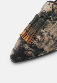 Chaussure à bout pointu avec motif floral multicolore et glands en cuir superposés en noir, olive et beige sur le dessus.