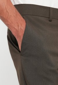 Dark green trousers with a smooth texture, showing a hand in the side pocket and a fitted waistband. Tailored design detail visible.