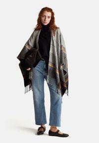 Szary i czarny ponczo w kratę z frędzlami, noszone na czarnym swetrze. Połączone z jasnoniebieskimi prostymi dżinsami i czarnymi płaskimi butami.