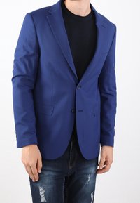 Blazer blu con texture, chiusura a un bottone, revers a lancia e tasche laterali, abbinato a una camicia nera e jeans strappati.