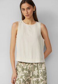 Crème mouwloos topje met een ronde halslijn, voorzien van een relaxed fit en subtiele stiksels. Gecombineerd met groene broeken met bloemenpatroon.