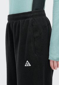Μαύρο φανελένιο παντελόνι φόρμας με ελαστική μέση και πλευρικές τσέπες, που διαθέτει λευκό τριγωνικό λογότυπο στο κάτω μέρος του ποδιού.