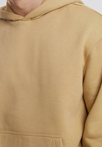 Osoba oblečená v béžové mikině s kapucí a přední kapsou, zobrazuje horní část trupu a část krku.