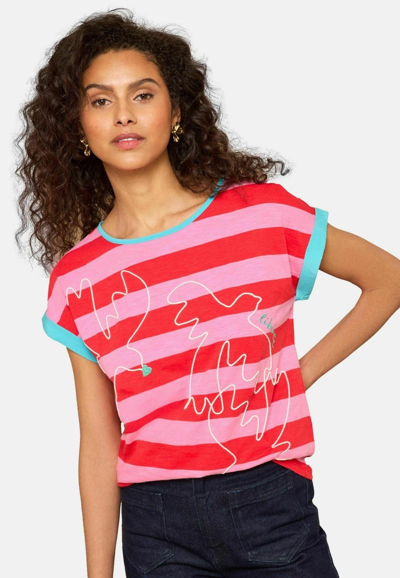 Kurdidega naine, kes kannab punaseid ja roosasid triipse ning türkiissinise äärisega särki, millel on valgete lindude joonistused, koos tumedate pükstega.