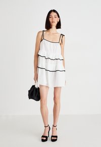 Robe blanche à volants avec des bordures noires, bretelles spaghetti et volants superposés. Mannequin tenant un sac noir et portant des sandales à talons hauts noires.