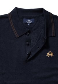 Laivastonsininen poolopaita, jossa on kaksi ruskeaa raitaa kauluksessa, kolme nappia ja kultakirjailtu logo, jossa on kaksi leijonaa.