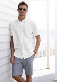 Camicia bianca a maniche corte con bottoni e taschino, abbinata a pantaloni corti grigio chiaro. Il modello è in piedi contro una parete bianca e indossa occhiali da sole.