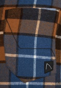 Kieszeń z tkaniny w kratę w kolorach brązowym, niebieskim, białym i czarnym, z przeszytymi krawędziami oraz małą czarną naszywką z białym abstrakcyjnym wzorem w dolnym rogu.