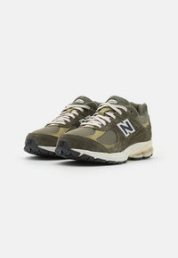 Ett par olivgröna och beige New Balance löparskor med texturiserat mesh, mockadetaljer, vita snören och sulor med svart mönstring på vit bakgrund.