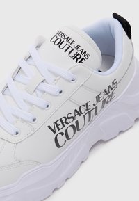 Baskets blanches en matériau synthétique lisse avec le texte noir 'Versace Jeans Couture', fermeture à lacets et semelle épaisse texturée.