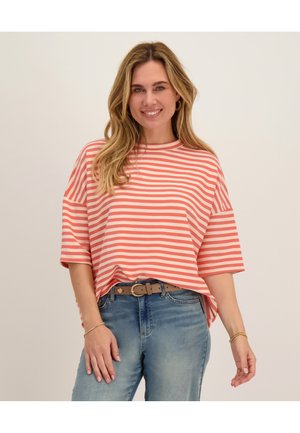 Vrouw lacht en draagt een koraalrode en wit gestreepte top met korte mouwen, blauwe spijkerbroek en een beige riem met gouden accessoires.