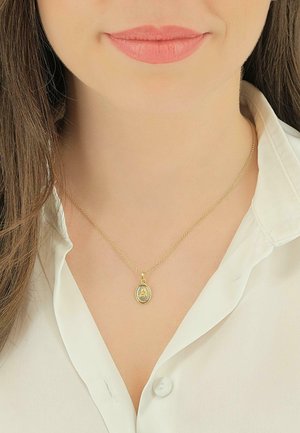 Collier en or avec une chaîne fine et un pendentif ovale présentant une figure détaillée. Porté sur une chemise blanche, mettant en valeur son design délicat.