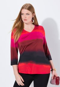 V-Ausschnitt, Dreiviertelarmoberteil mit einem Farbverlaufsmuster in lebhaftem Pink, Rot und Schwarz. Hergestellt aus weichem Stoff, ergänzt durch eine glänzende rote Handtasche.