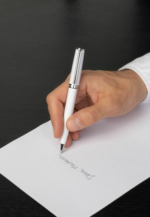 Weißer Stift mit metallischen Akzenten, der in einer Hand gehalten wird und auf einem weißen Blatt Papier schreibt. Der Hintergrund ist eine dunkle Holzoberfläche.