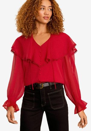 Rote Chiffon-Bluse mit einem gestuften Rüschendesign, V-Ausschnitt, Knopfleiste und langen transparenten Ärmeln mit elastischen Manschetten. Kombiniert mit schwarzen Hosen.