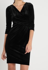 Svart sammetsinlindad klänning med trekvartslånga ärmar, v-ringning, rynk vid midjan och en figurnära silhuett. Slät, mjuk textur.