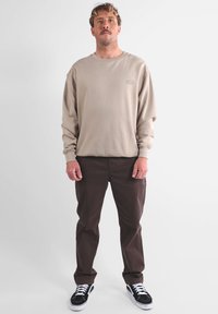 Chemise beige à manches longues avec col ras du cou, présentant un petit logo. Pantalons marron foncé et baskets noires avec lacets blancs. Design simple, coupe décontractée.