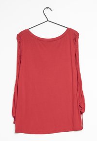 Červená košile s dlouhým rukávem a kulatým výstřihem, s nařasením na rukávech, vyrobená z jemné látky, zavěšená na čern ém háčku.