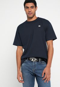 Μπλε ναυτικό βαμβακερό μπλουζάκι με άνετη εφαρμογή, κοντά μανίκια και μικρό διακοσμητικό λογότυπο· φορεμένο με τζιν και μαύρη ζώνη με ασημένια αγκράφα.