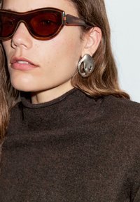 Brun polokragsweater, transparenta sköldpaddsglasögon med mörka linser och stora skulpturala örhängen i silver. Släta texturer och modern design.